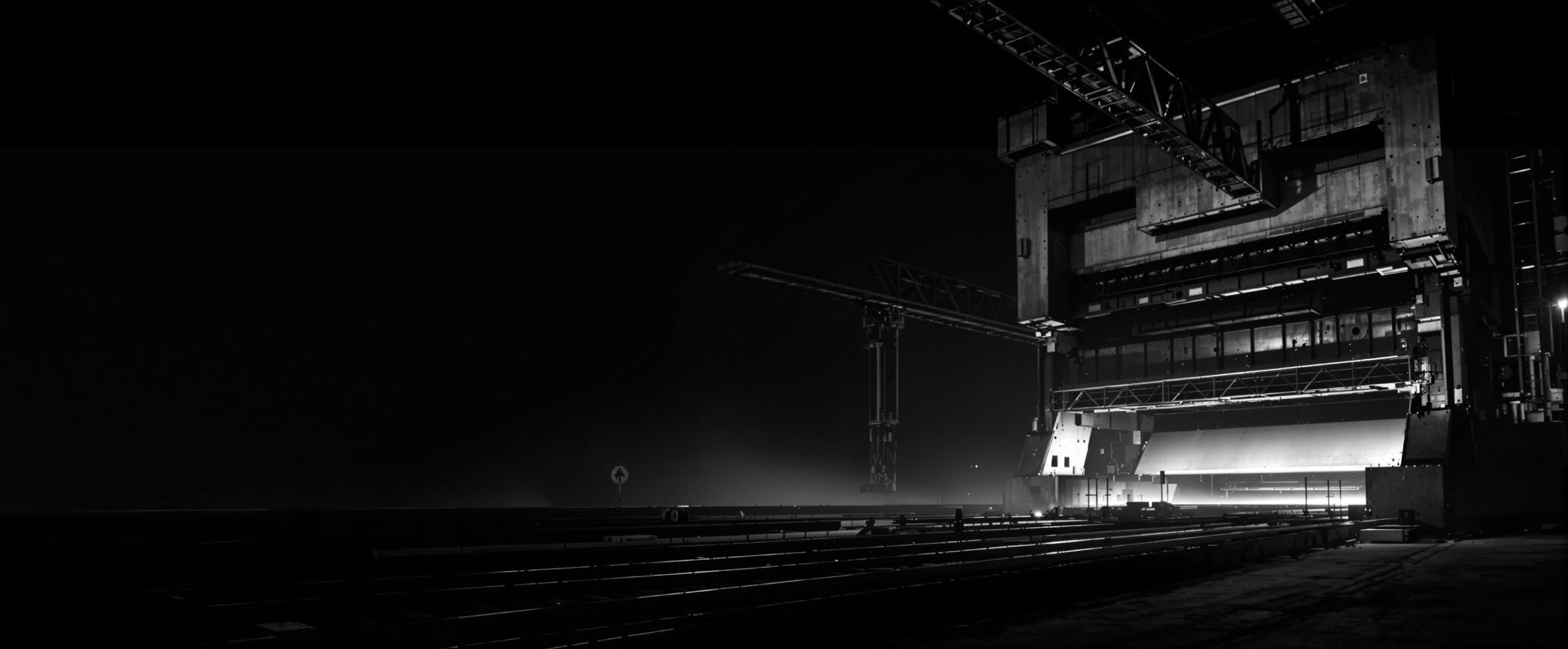 Heavy machinery plant at night: a steel press and the rail of an overhead crane lit by hard work light, with the hall dark around them.